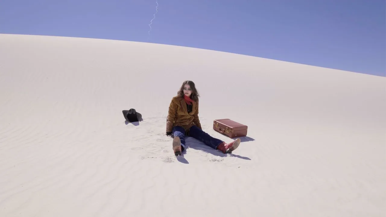 Person sitting beside a suitcase on a pale sand dune.