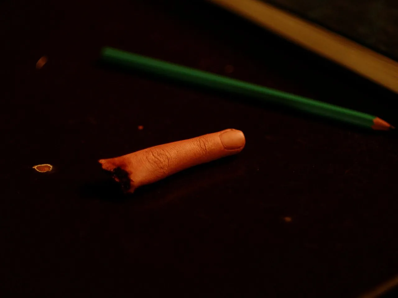 Severed-finger prop on a dark surface beside a green pencil.